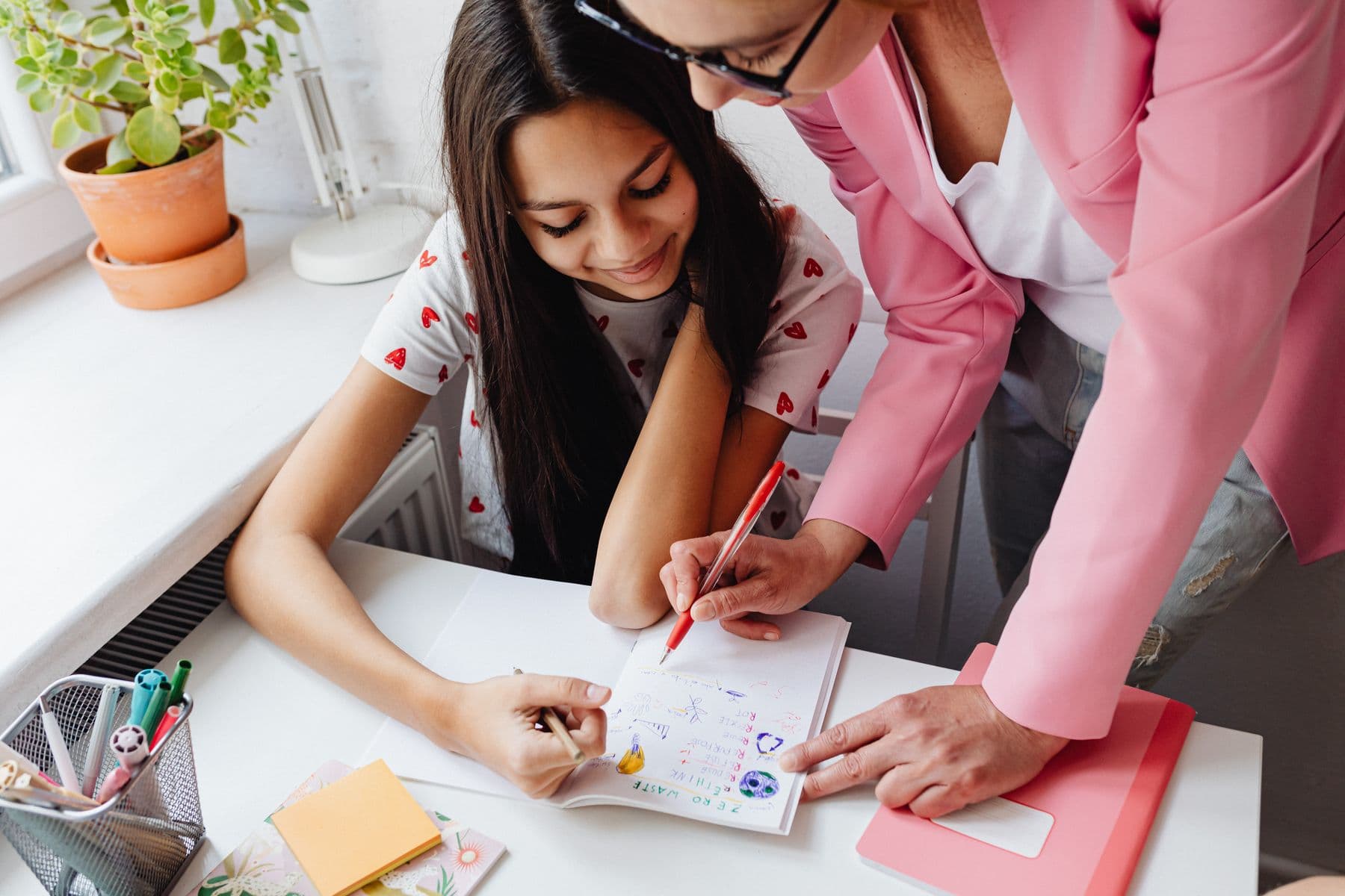 An adult leaning over a desk to guide a young person working in a notebook.