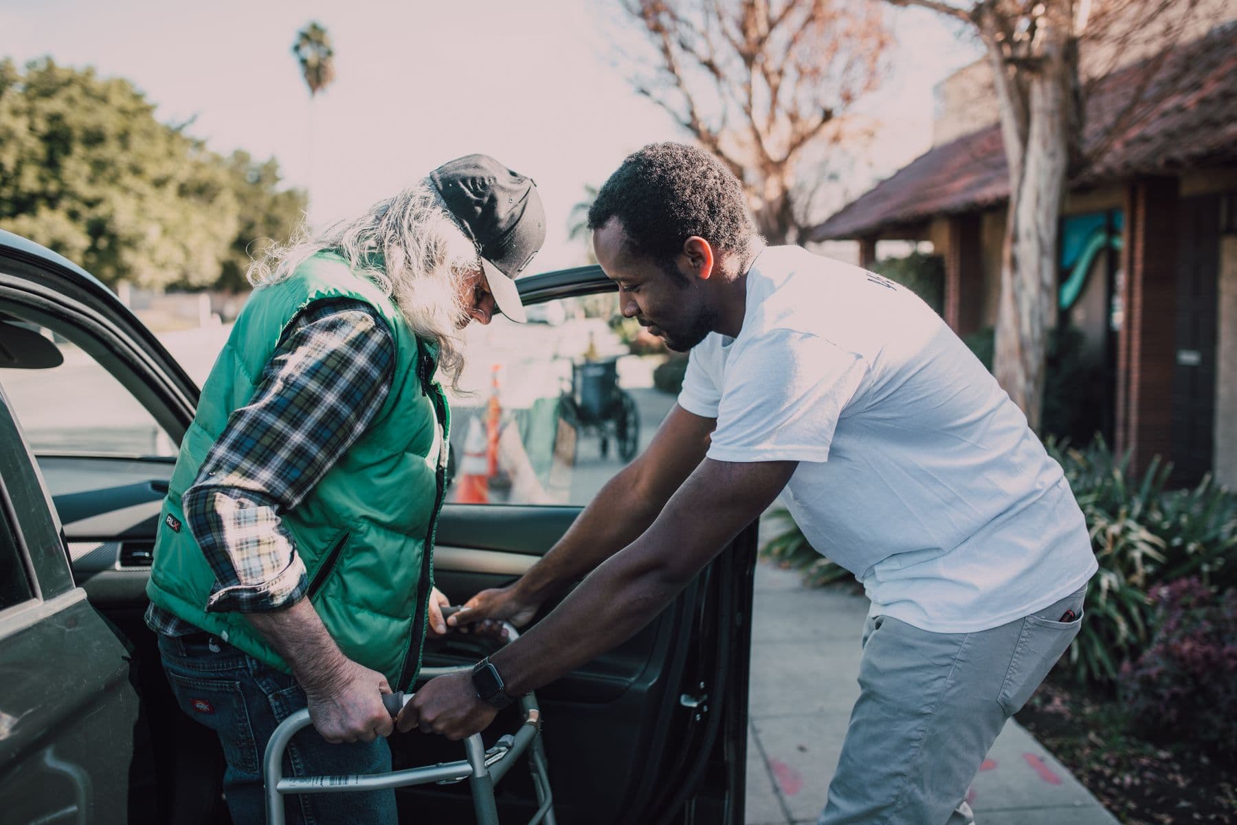 A person steadying an older adult with a walking frame beside an open car door.