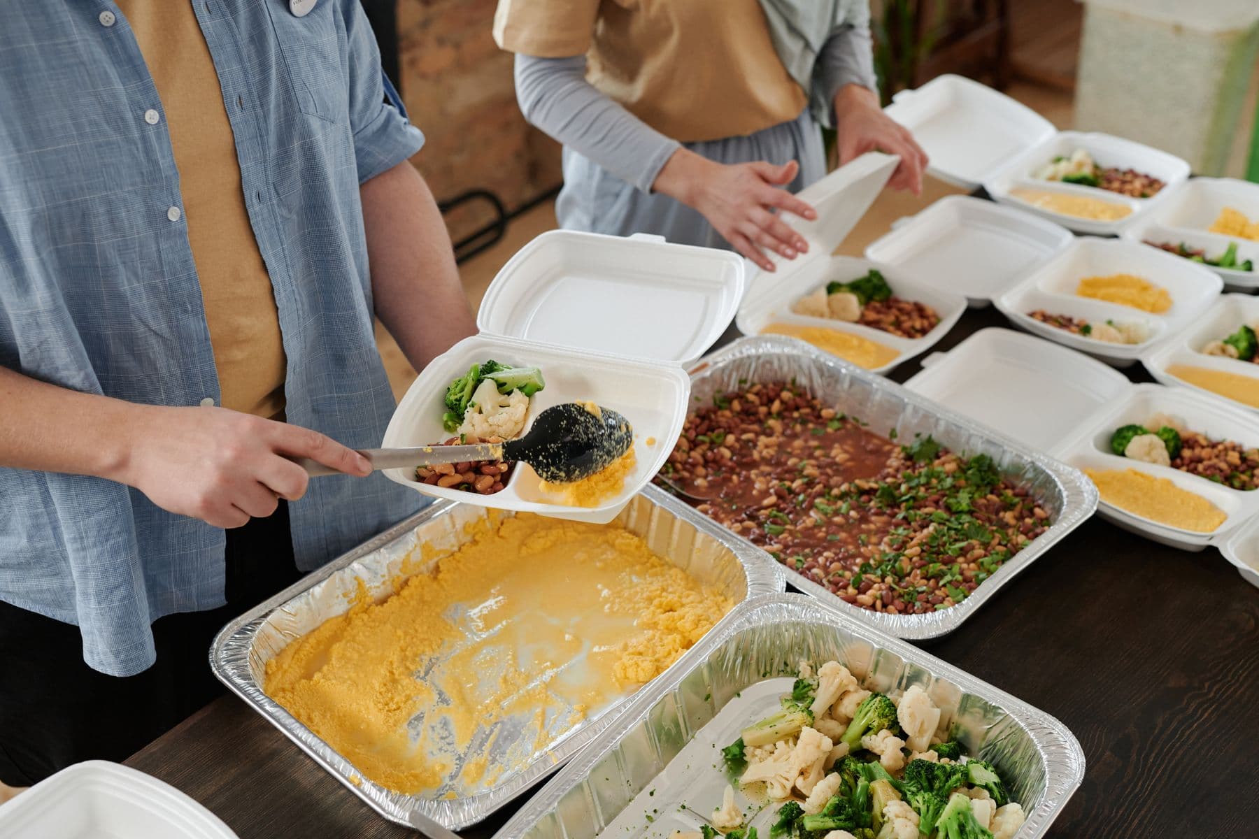 Hands spooning cooked food into takeaway containers from foil trays.